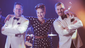 A woman wearing a polka-dot dress with a 40s hairstyle, is flanked by two men in white tuxedos - her arms are across their shoulders. Both men have their arms crossed and they're all smiling widely for the camera. There is a drumkit behind them.