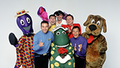 The four original Wiggles - Red, Yellow, Purple and Blue - stand in a neutral photography studio a group, smiling widely and doing finger-gun gestures with their hands. Captain Feathersword, Dorothy the Dinosaur, Henry the Octopus and Wags the Dog stand with them.