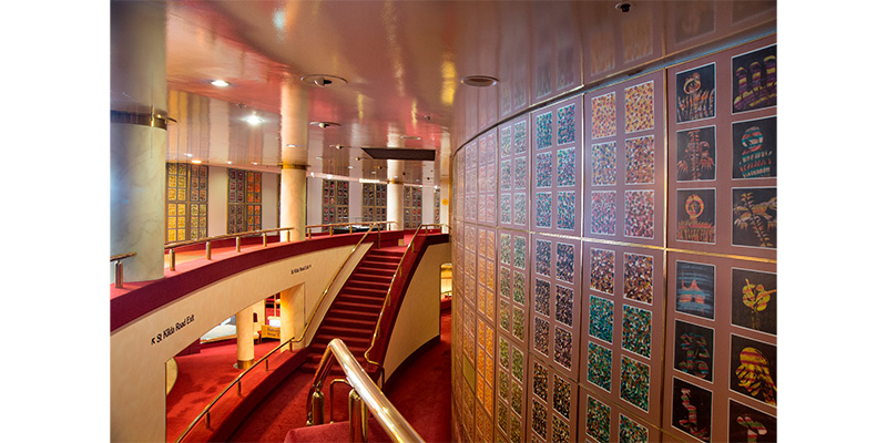 A view of part of 'Paradise Garden' installed on a curved wall in the State Theatre foyer of Arts Centre Melbourne. In the background you can see two levels of the foyer and a set of stairs, and then more panels of 'Paradise Lost' installed on the far wall.