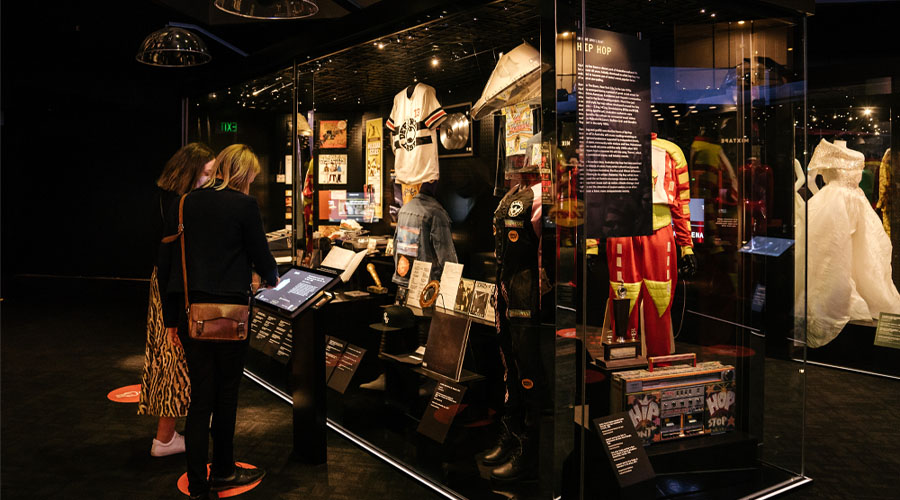 Two women enjoy the digital displays within the Australian Music Vault. In front of them is a touch-screen, and behind that is a large glass cabinet filled with costumes and music memorabilia.