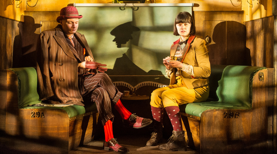 Two people, dressed on old-time clothes, are sitting in what looks like a train carriage. They are apprehensive and everything is a little bit surreal.