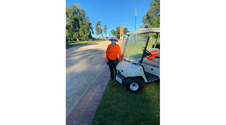 An Arts Centre Melbourne team member dressed in a hi-vis jacket, black trousers and a wide-brimmed hat, and wearing a mask, stands next to a small white buggy. Next to them is a tan path and on either side of the path are grass and trees.