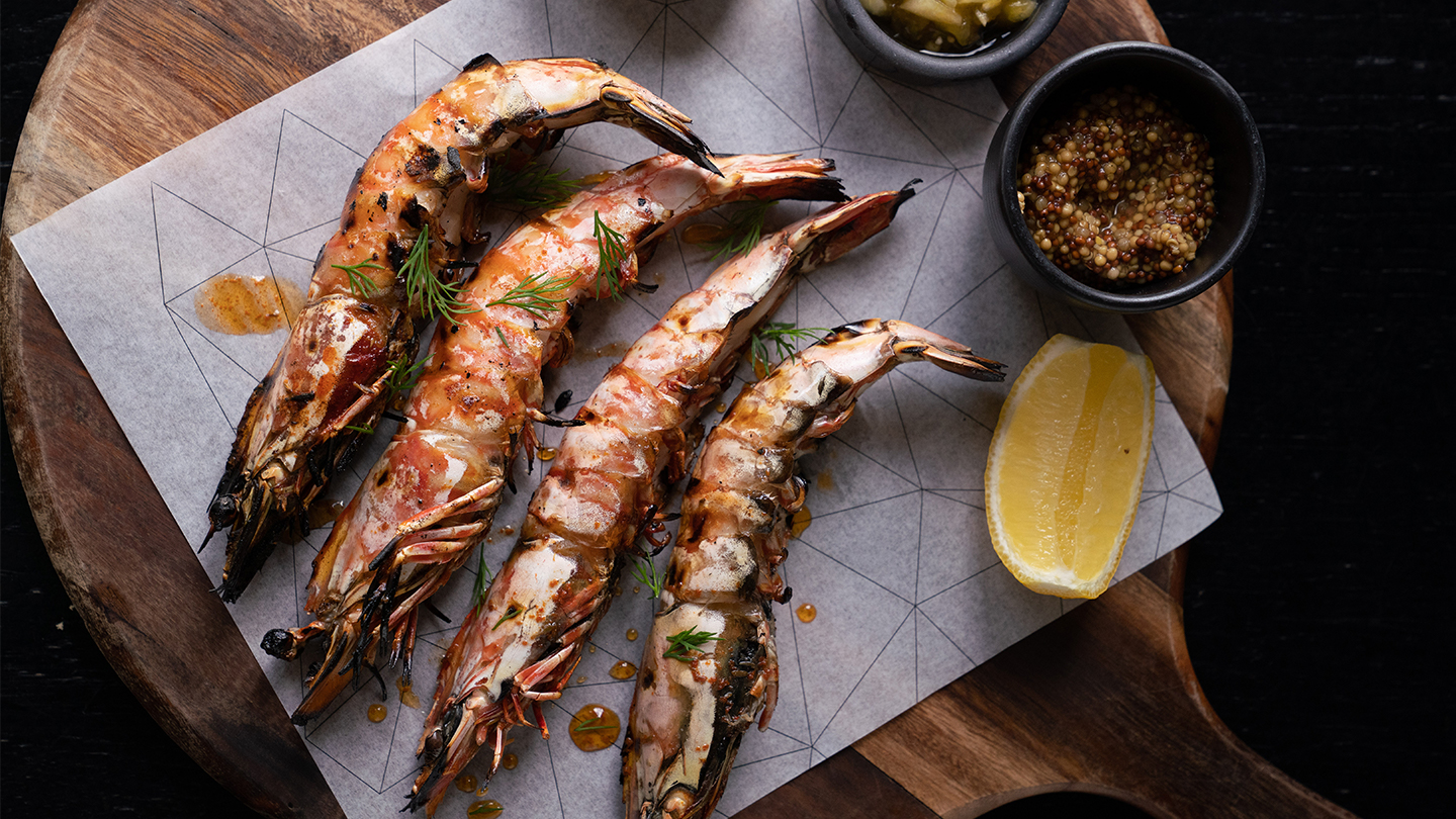 Four chargrilled prawns on a wooden board with a fresh slice of lemon.