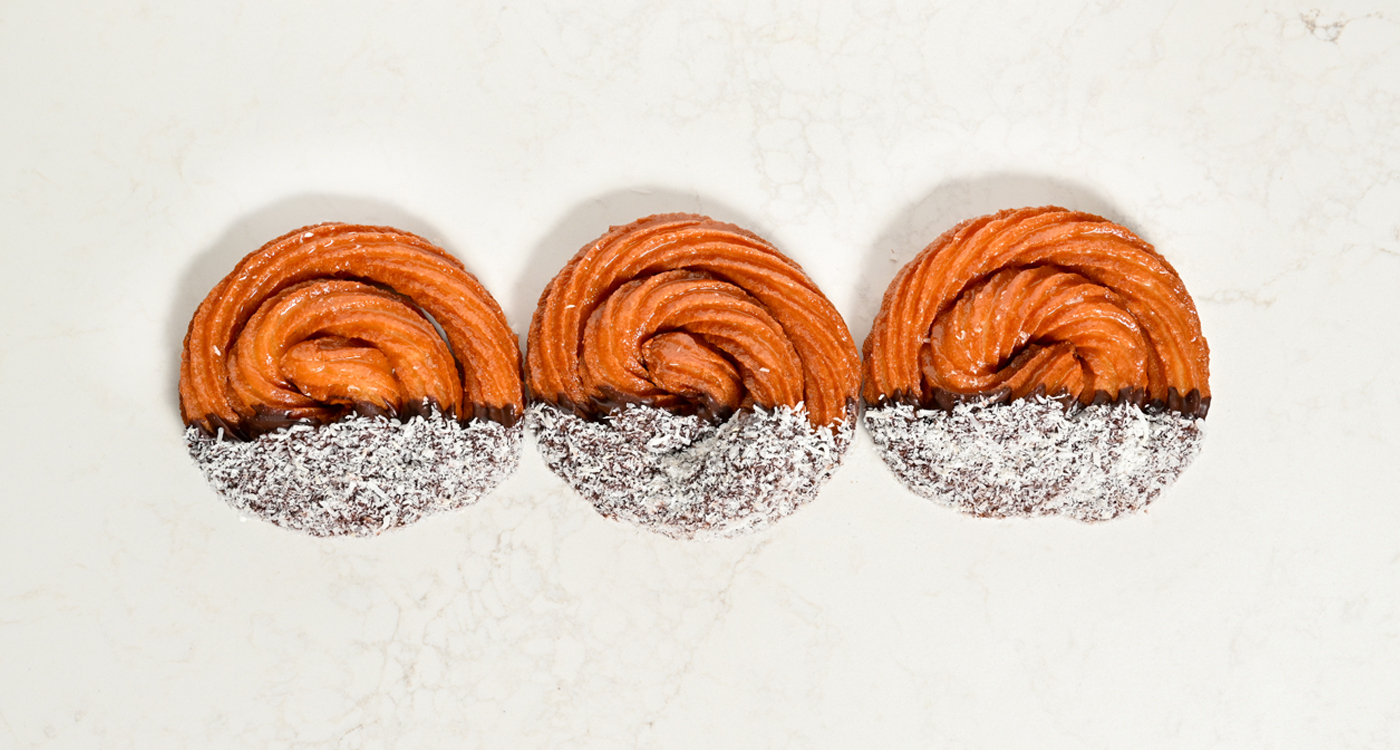 A flat lay style image of three Turkish donuts in a spiral shape, with half covered in a chocolate sauce and coconut.