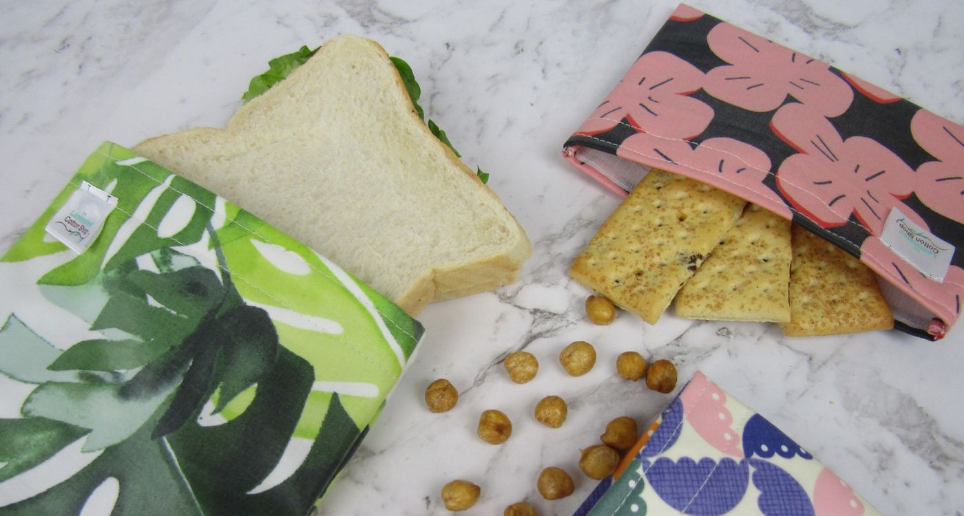 A collection of three patterned pouches, with a sandwich, crackers and chickpeas inside. 