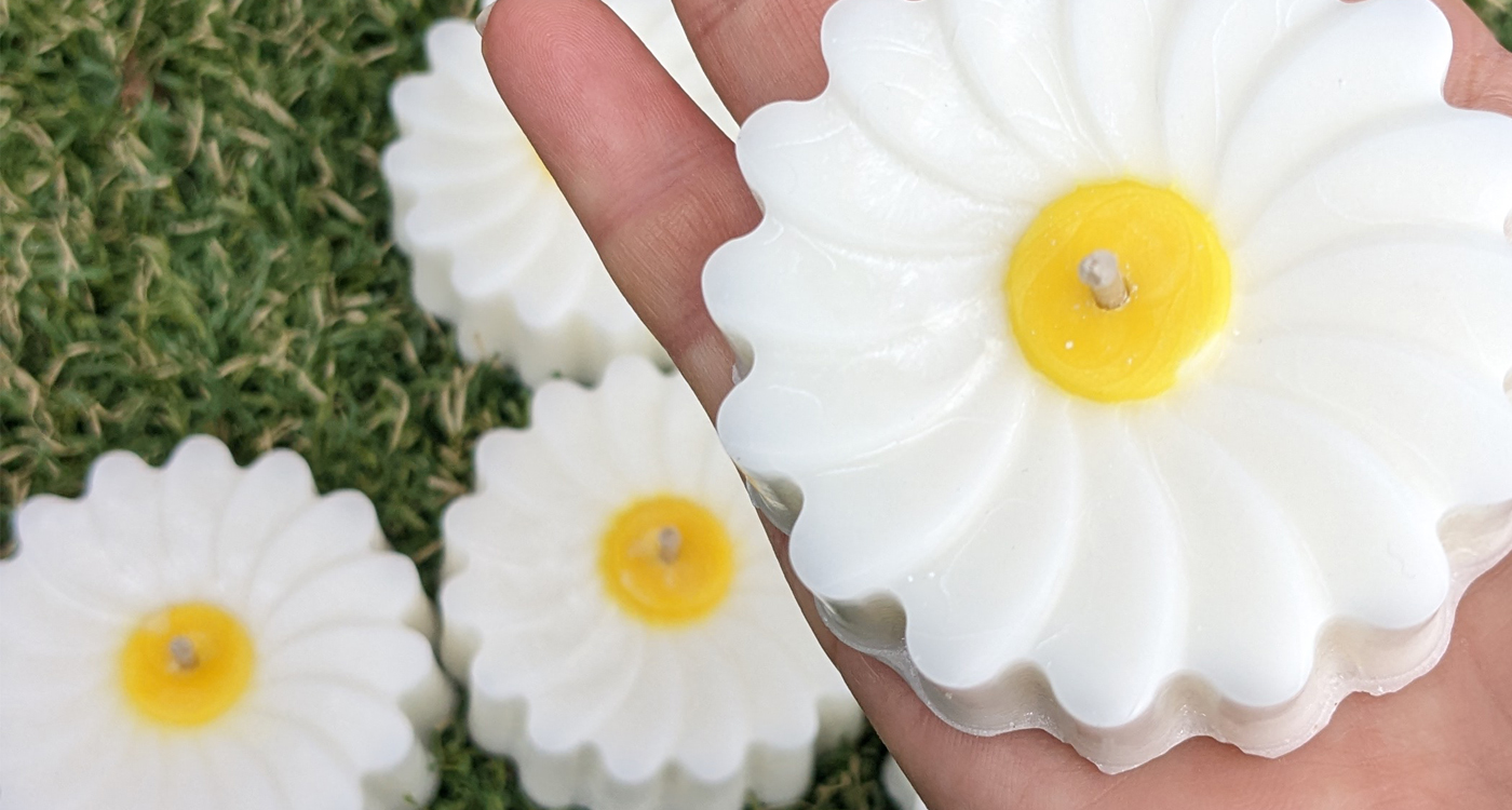 A top down image of a person holding a flower candle with white petals and yellow centre and wick. There are more candles in the background slightly out of focus. 