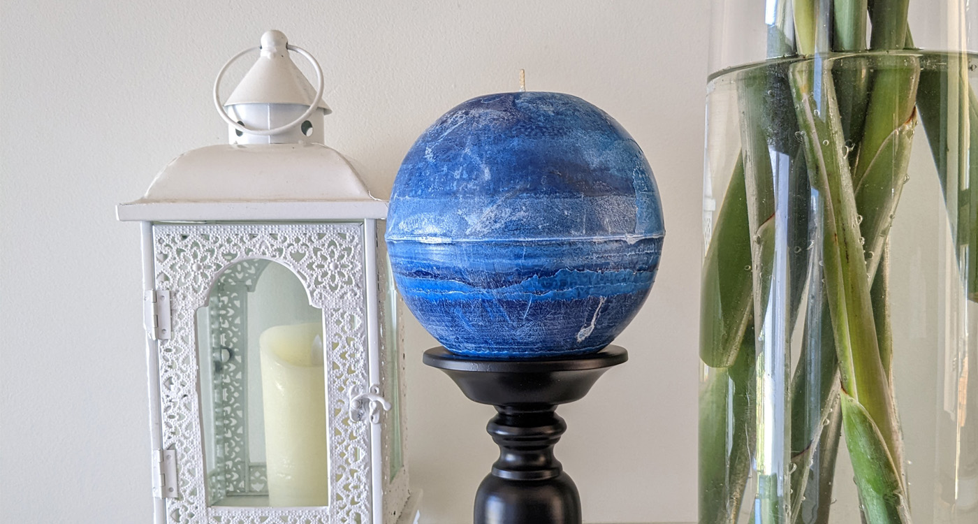 A side image of a large candle in a white lantern box, a blue circular candle on a black stand and the bottom of a vase with stems and water. 