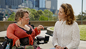 Caroline Bowditch, a woman with short asymmetrical blonde hair wearing a black top with a burnt orange overshirt, and Claire Spencer, a woman with curly brown hair wearing a white shirt, look at each other while talking. They are outside on the podium level of Southbank, Melbourne, and in the background is blue sky, trees and the Melbourne city skyline.